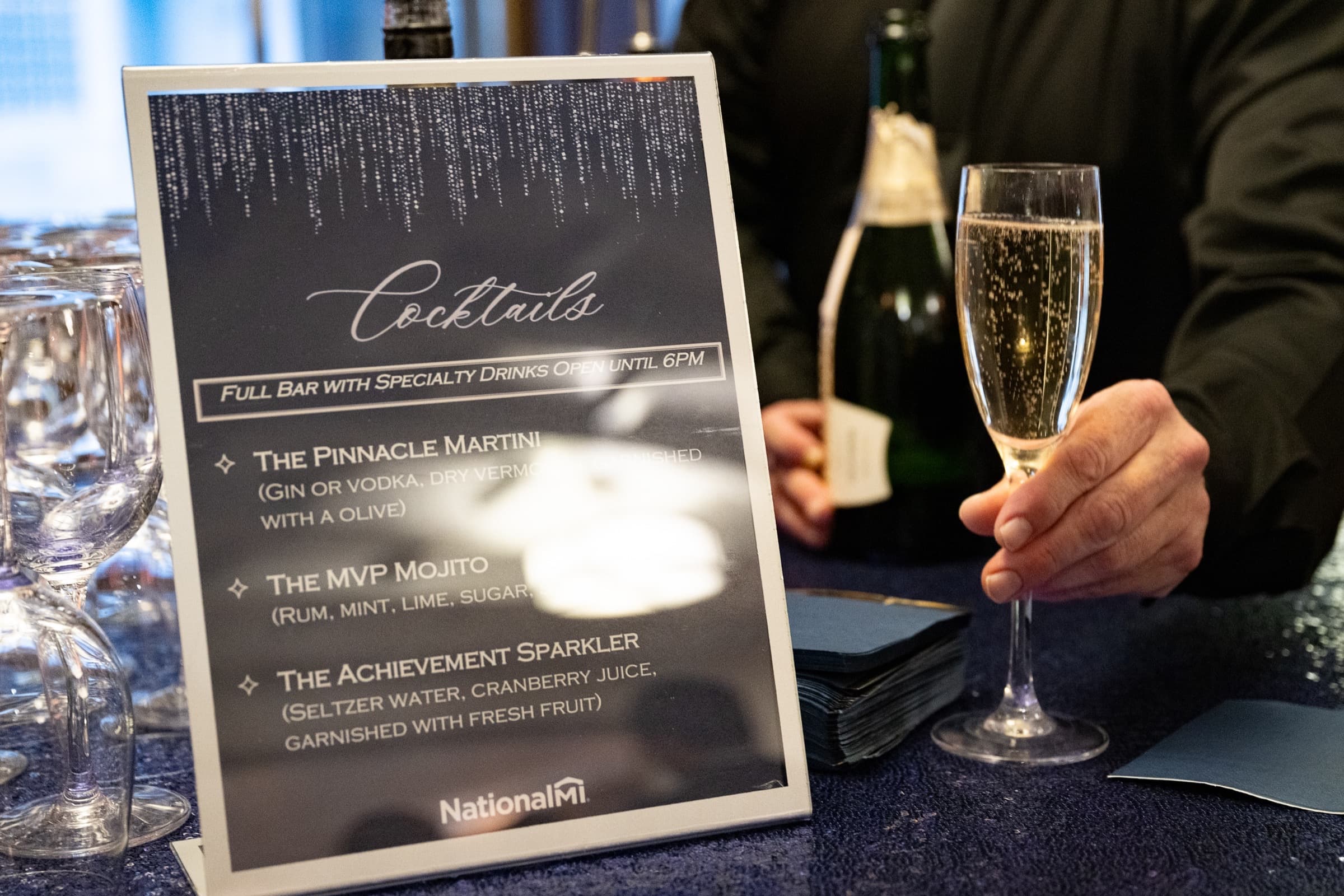Cocktail menu card beside a guest holding a champagne flute at a reception