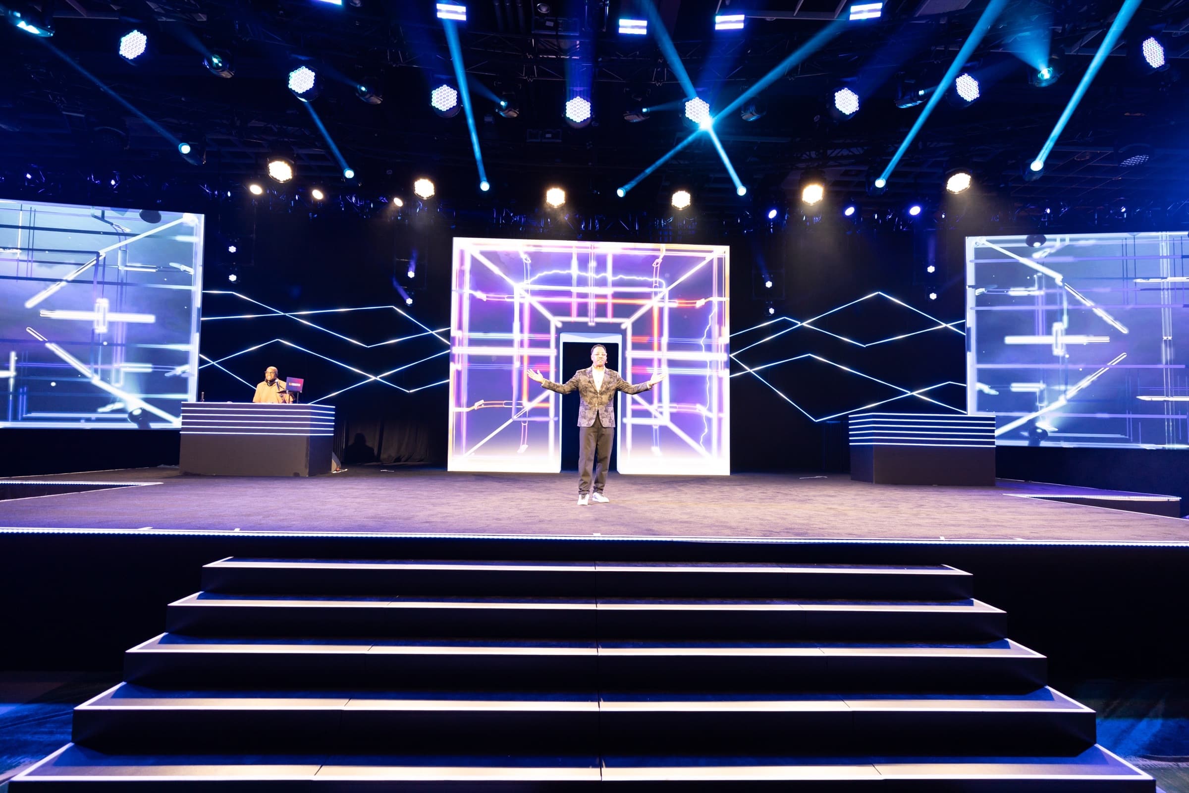 Single speaker on a wide, brightly lit general session stage with geometric LED walls
