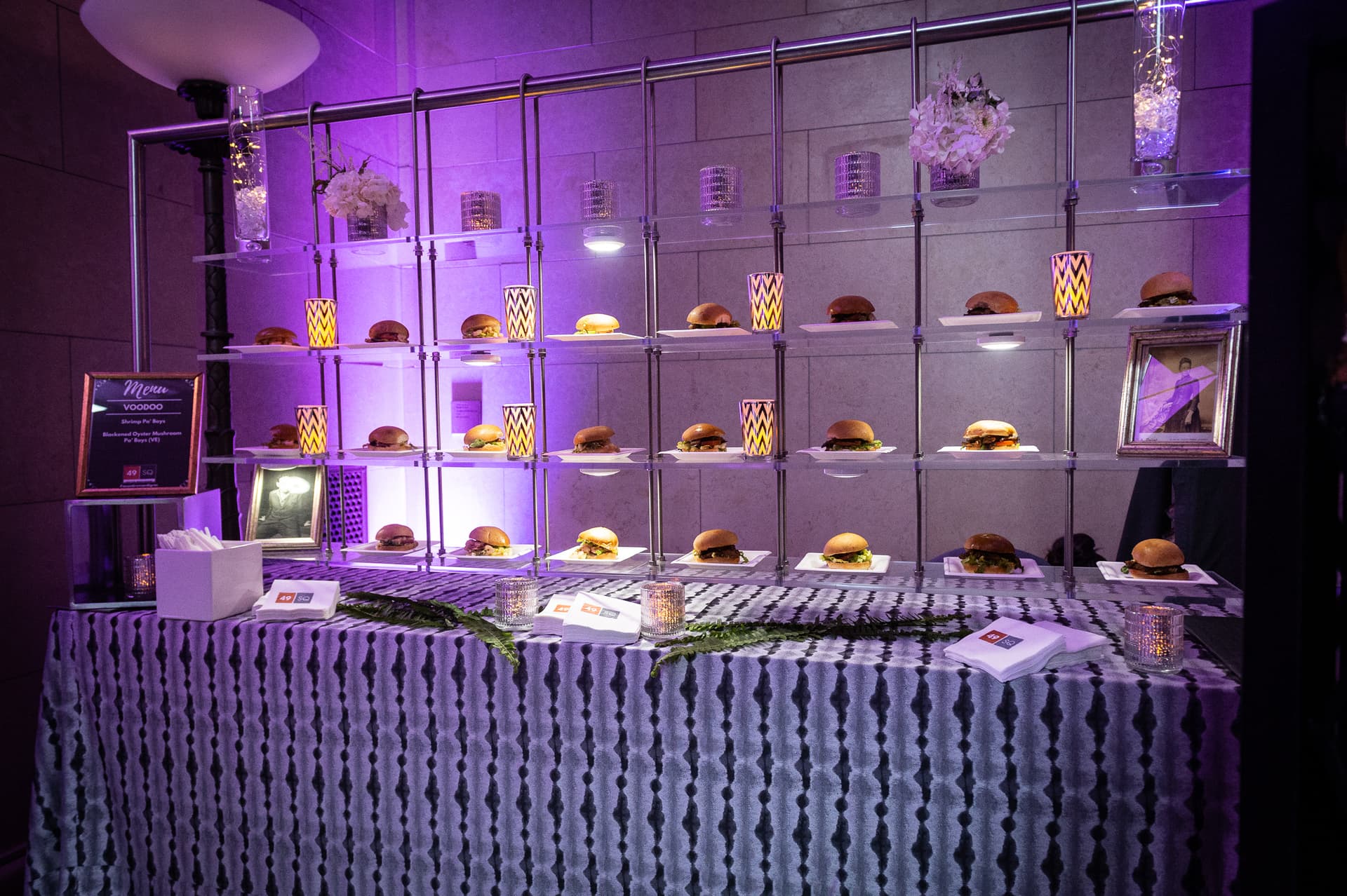 Dramatic purple-lit display wall showcasing gourmet burgers on illuminated shelving with decorative floral arrangements