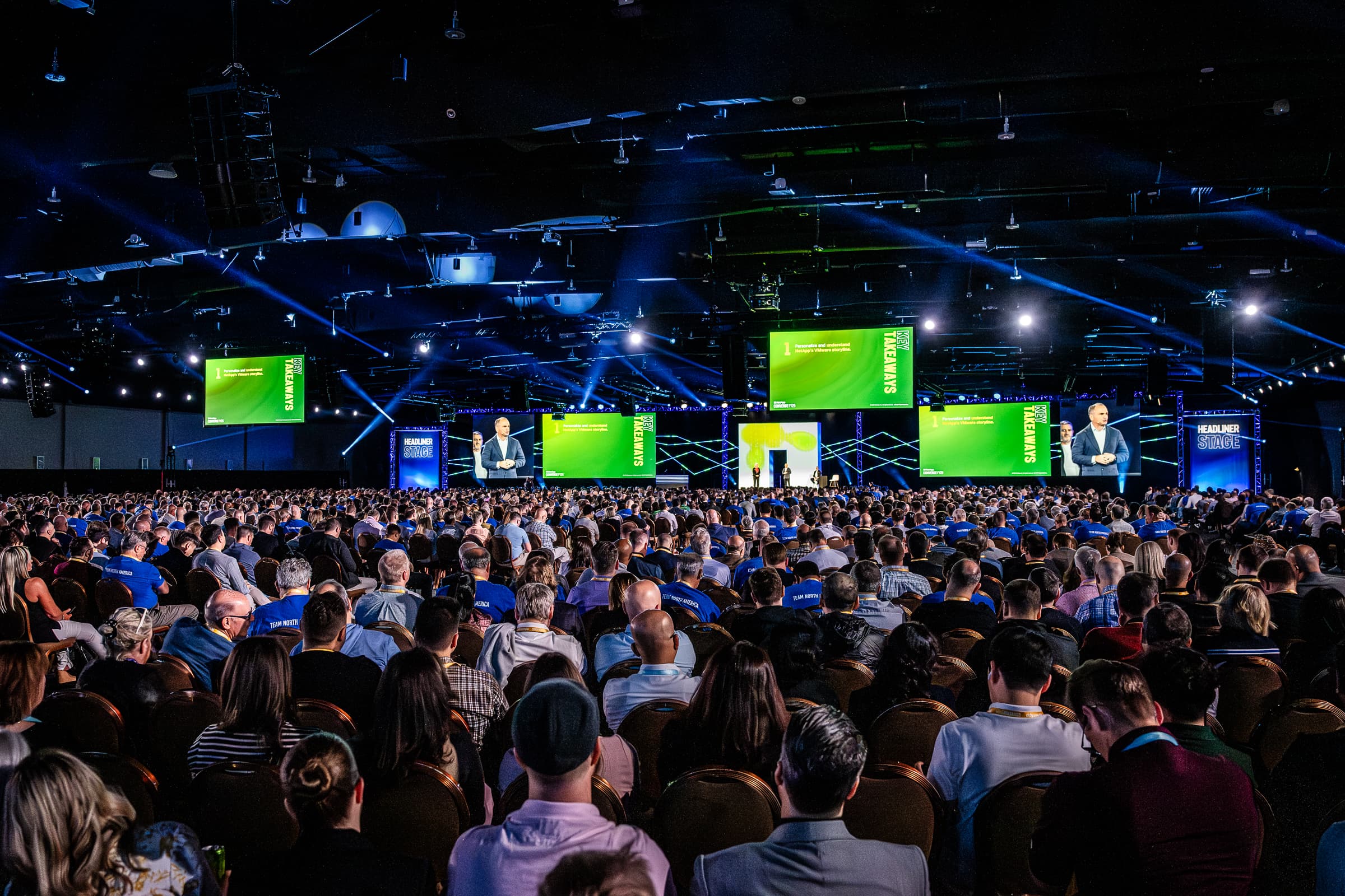 Full auditorium audience facing a bank of green-lit stage screens
