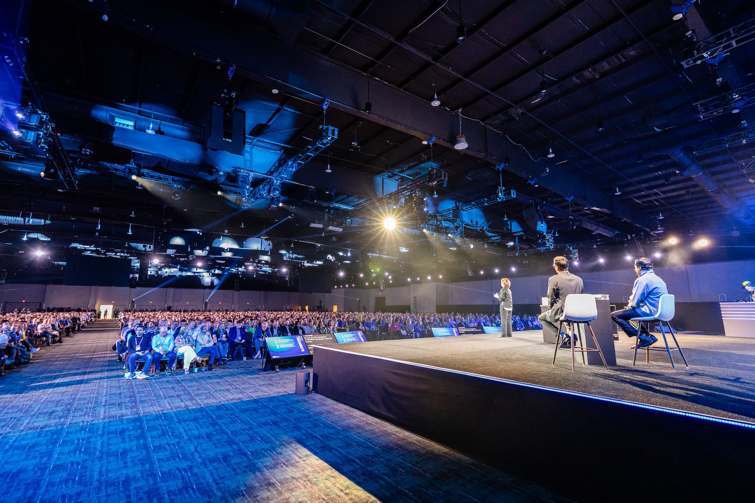 Three speakers on a runway stage in front of a full general-session audience