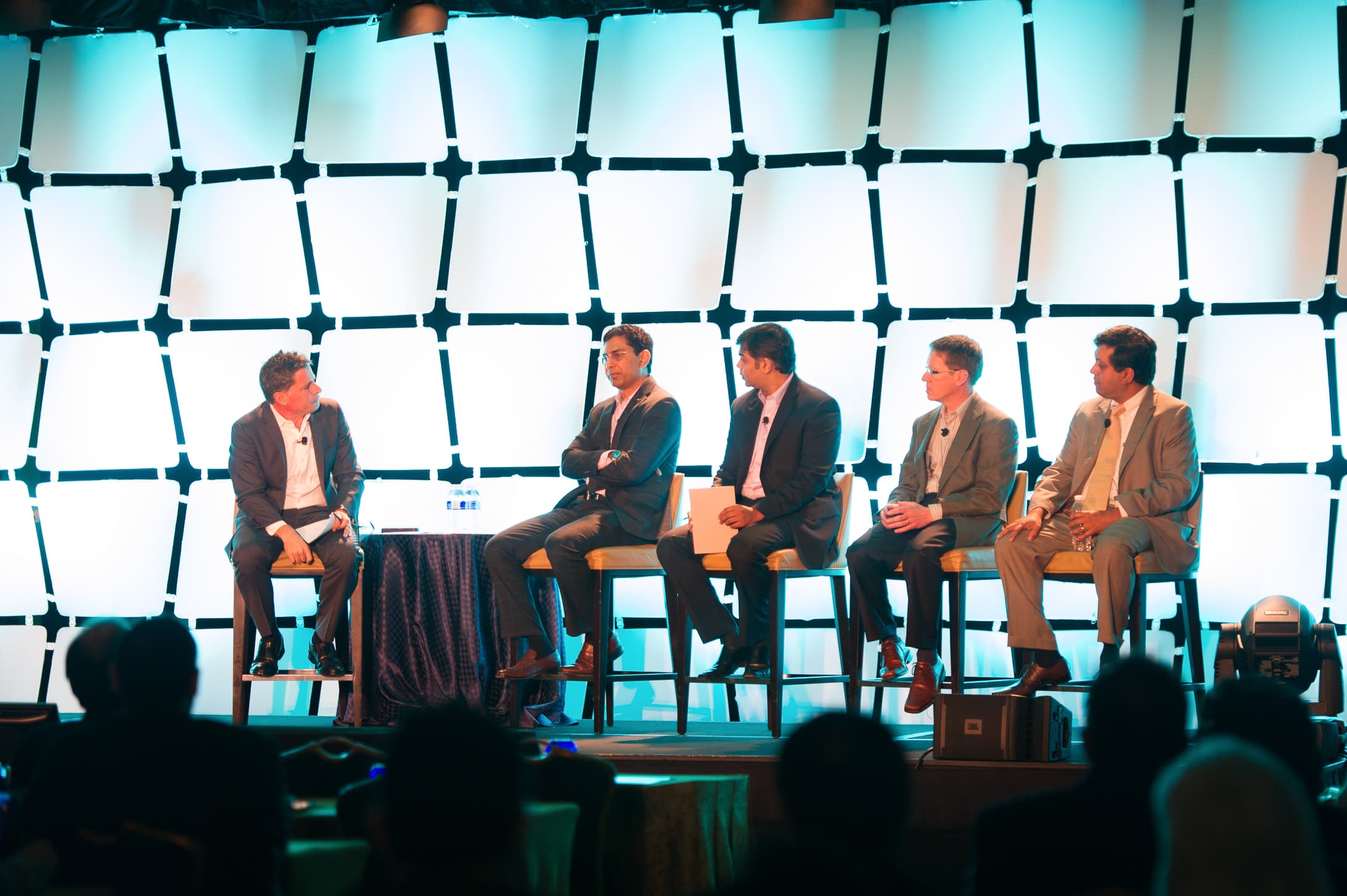 Six panellists seated on stage against an illuminated lattice backdrop