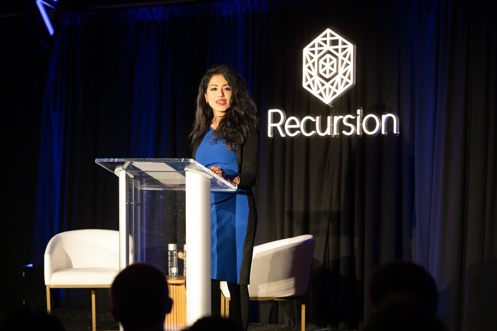 Speaker at a clear lectern beside illuminated Recursion branding at the San Francisco Mint