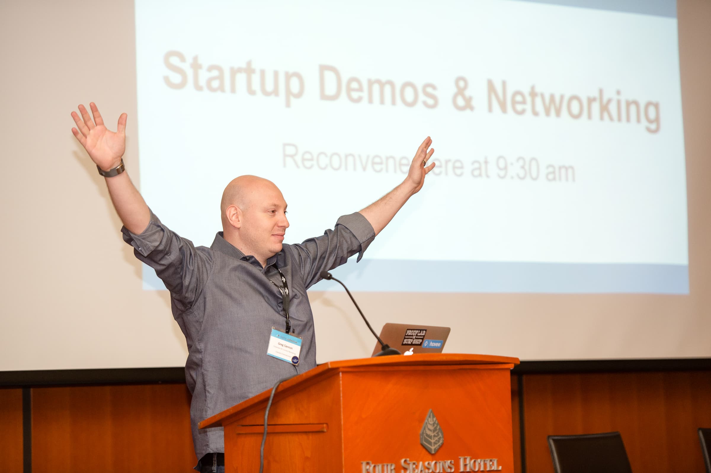 Speaker at a lectern with arms raised beside an agenda slide