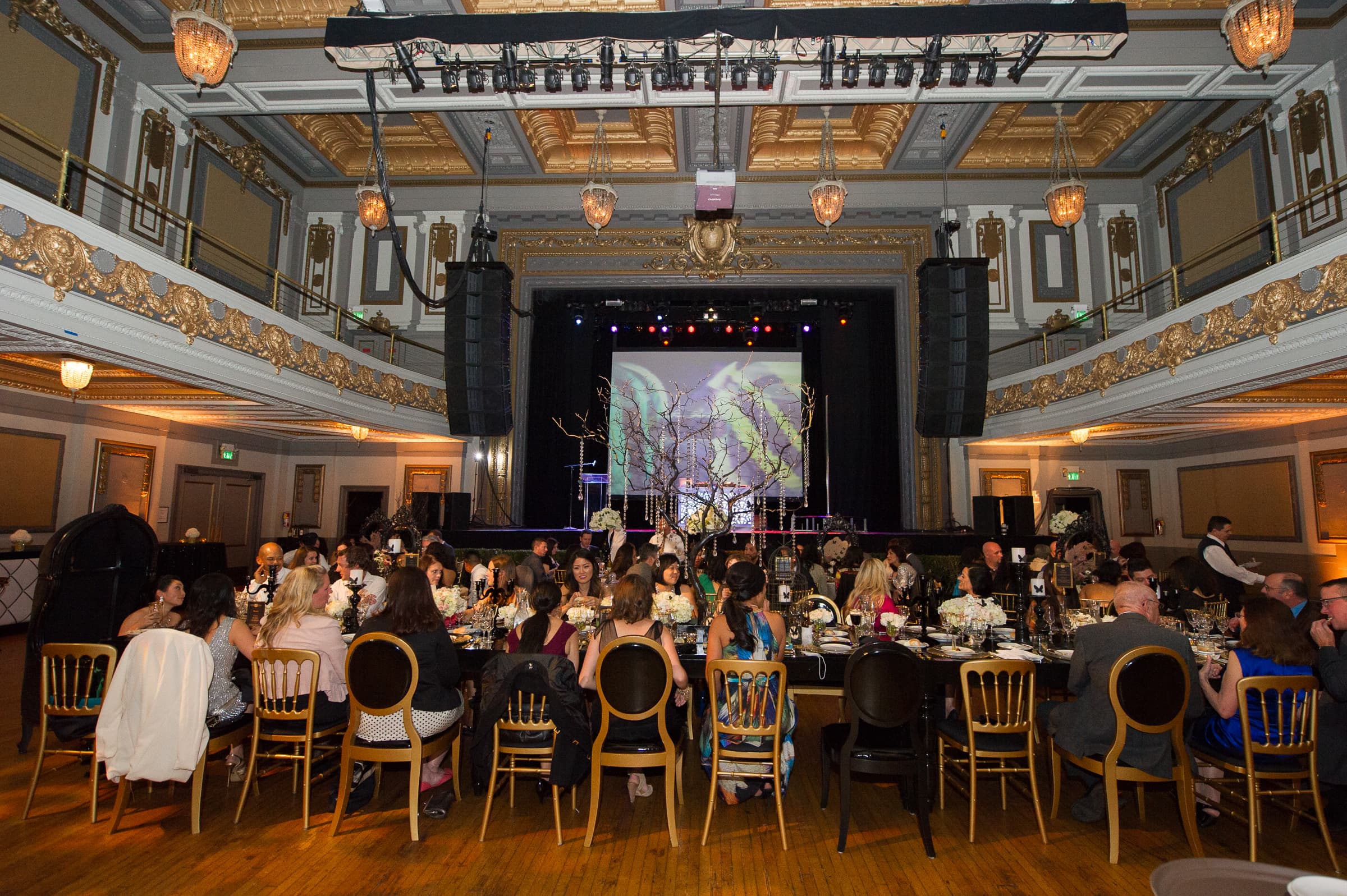 Gilded ballroom with guests seated at rounds facing a stage