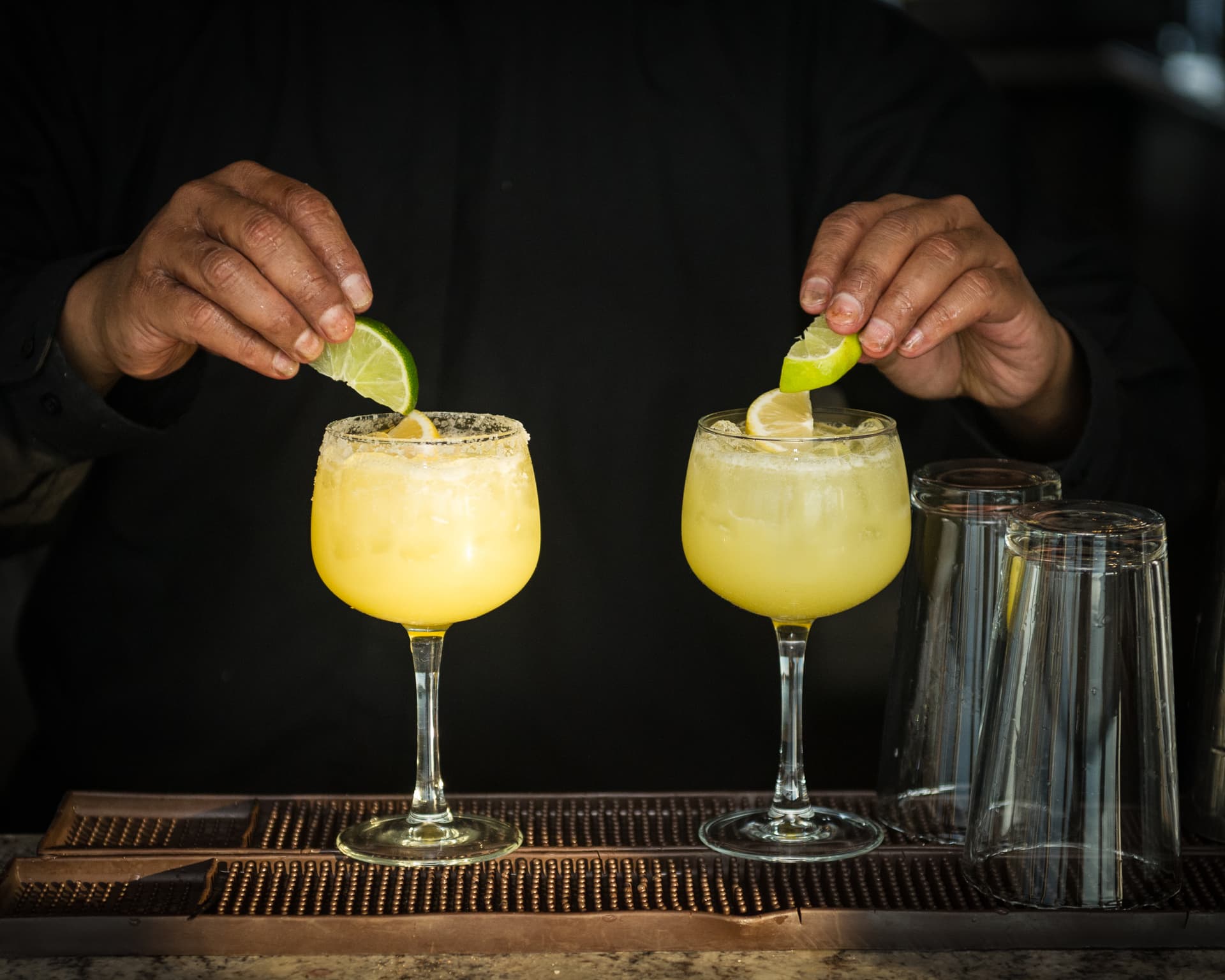 Bartender garnishing golden margarita-style cocktails with lime wedges on copper bar mat