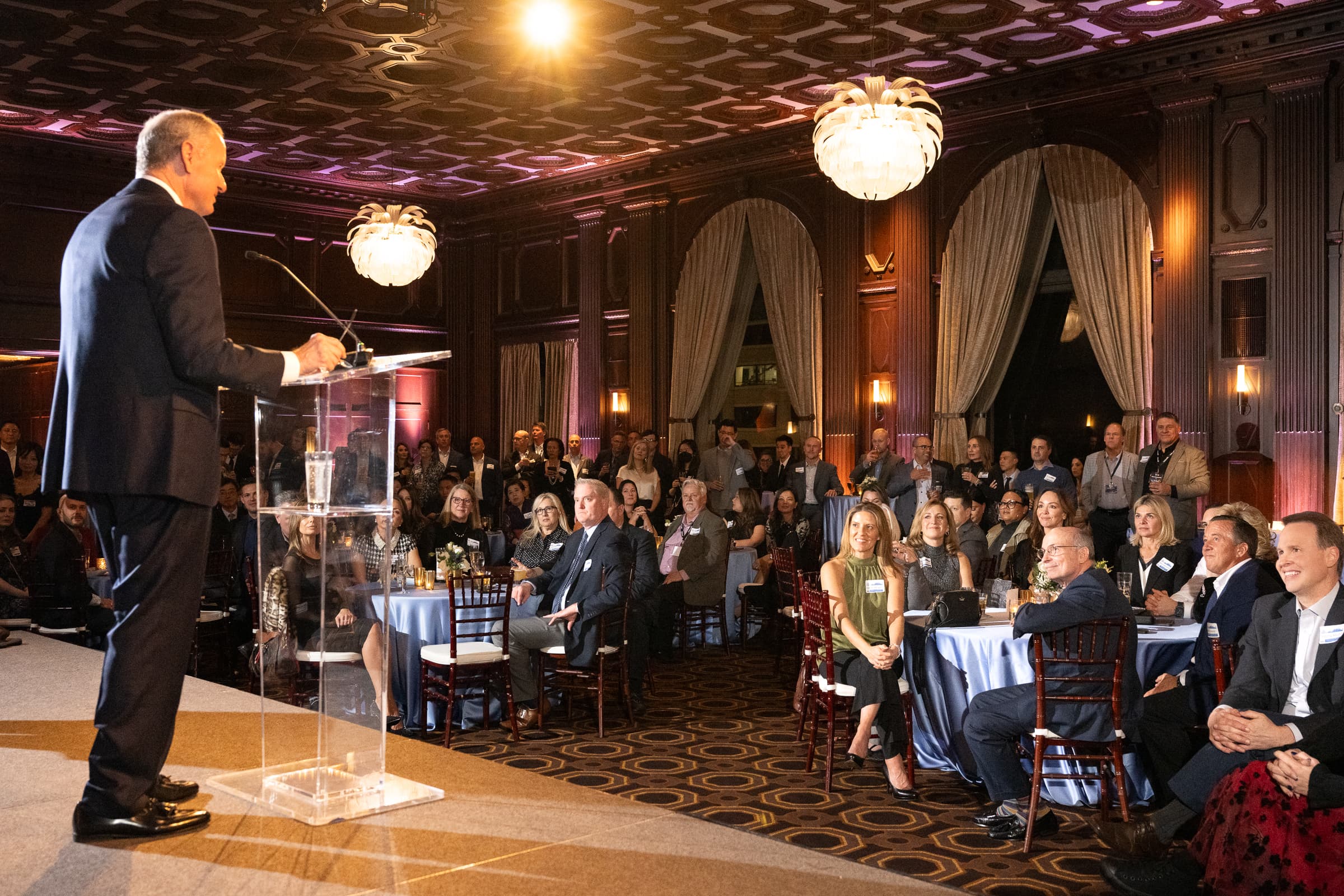Host giving remarks from a lectern to a full ballroom