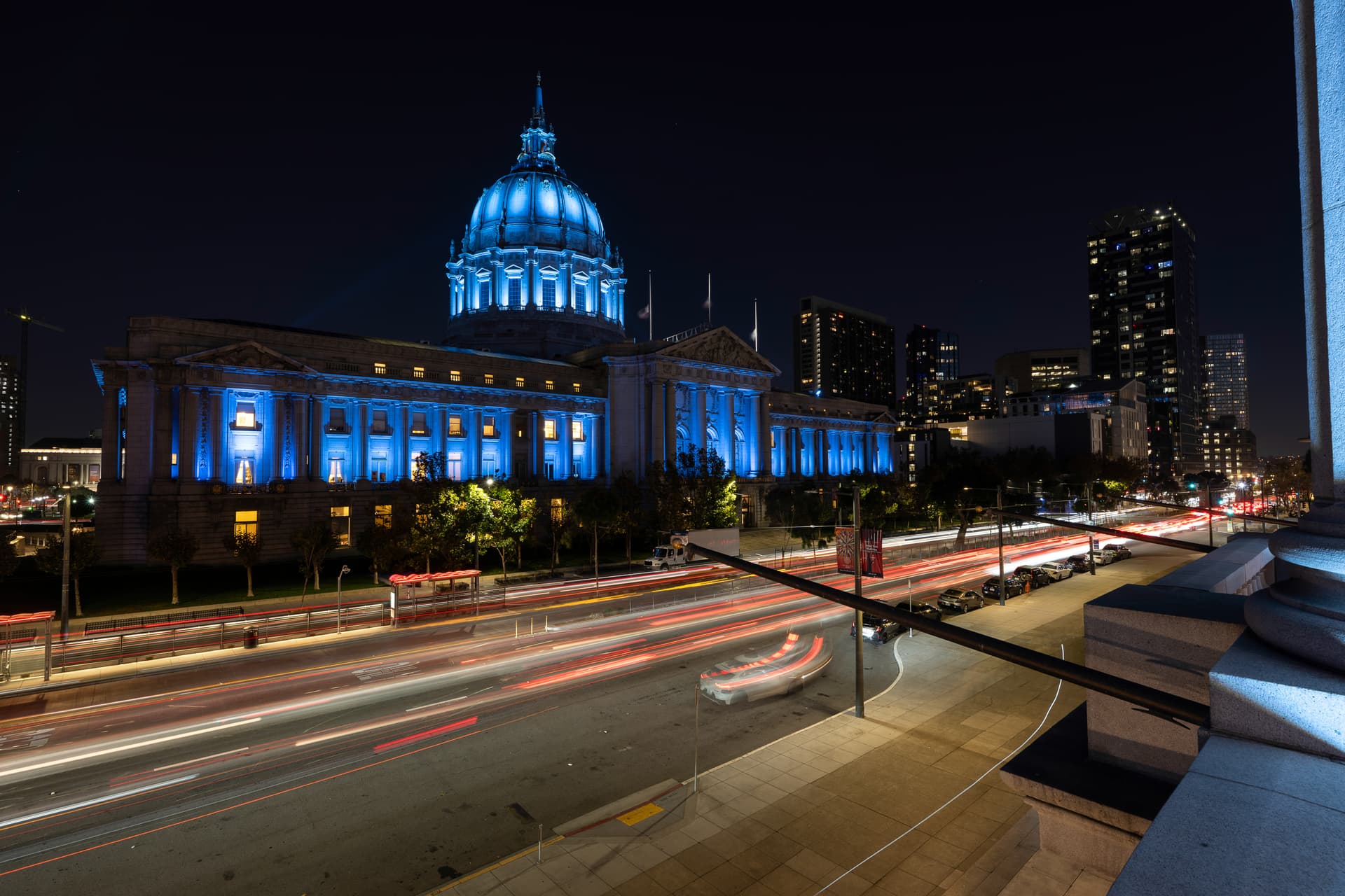 San Francisco City Hall illuminated blue at night with traffic light trails, viewed from the War Memorial