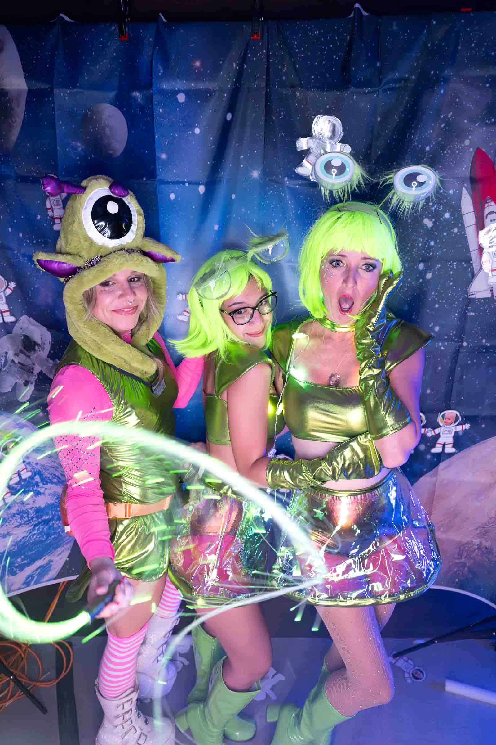 Three performers in elaborate neon alien costumes with glowing accessories posing against a starry space-themed backdrop