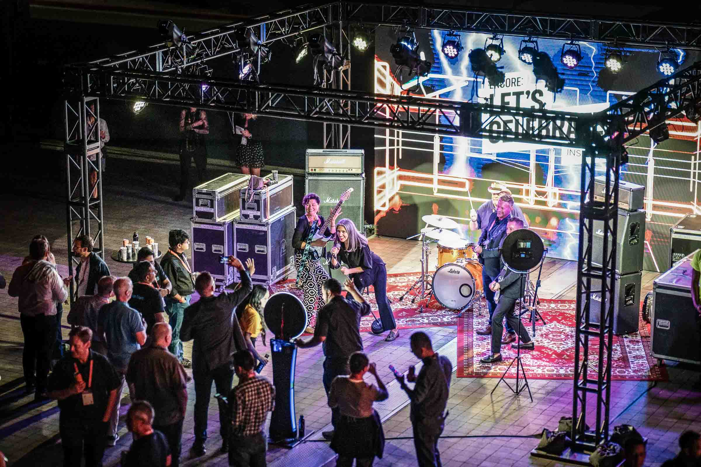 Live rock band performs on outdoor truss stage with purple lighting while crowd watches