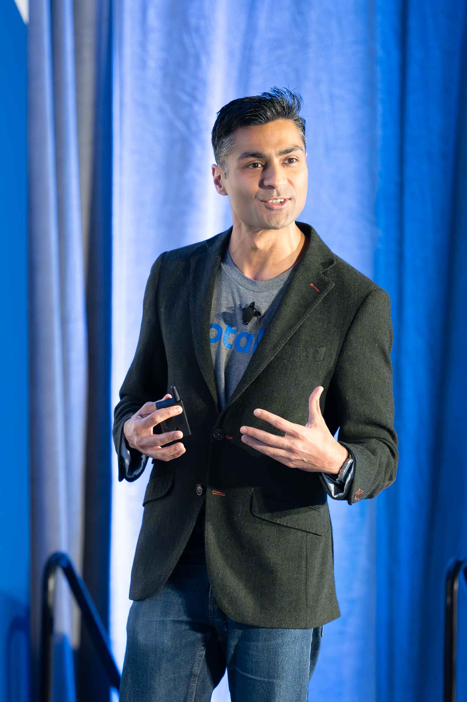 Man speaking on stage against blue draped lighting backdrop wearing business casual attire