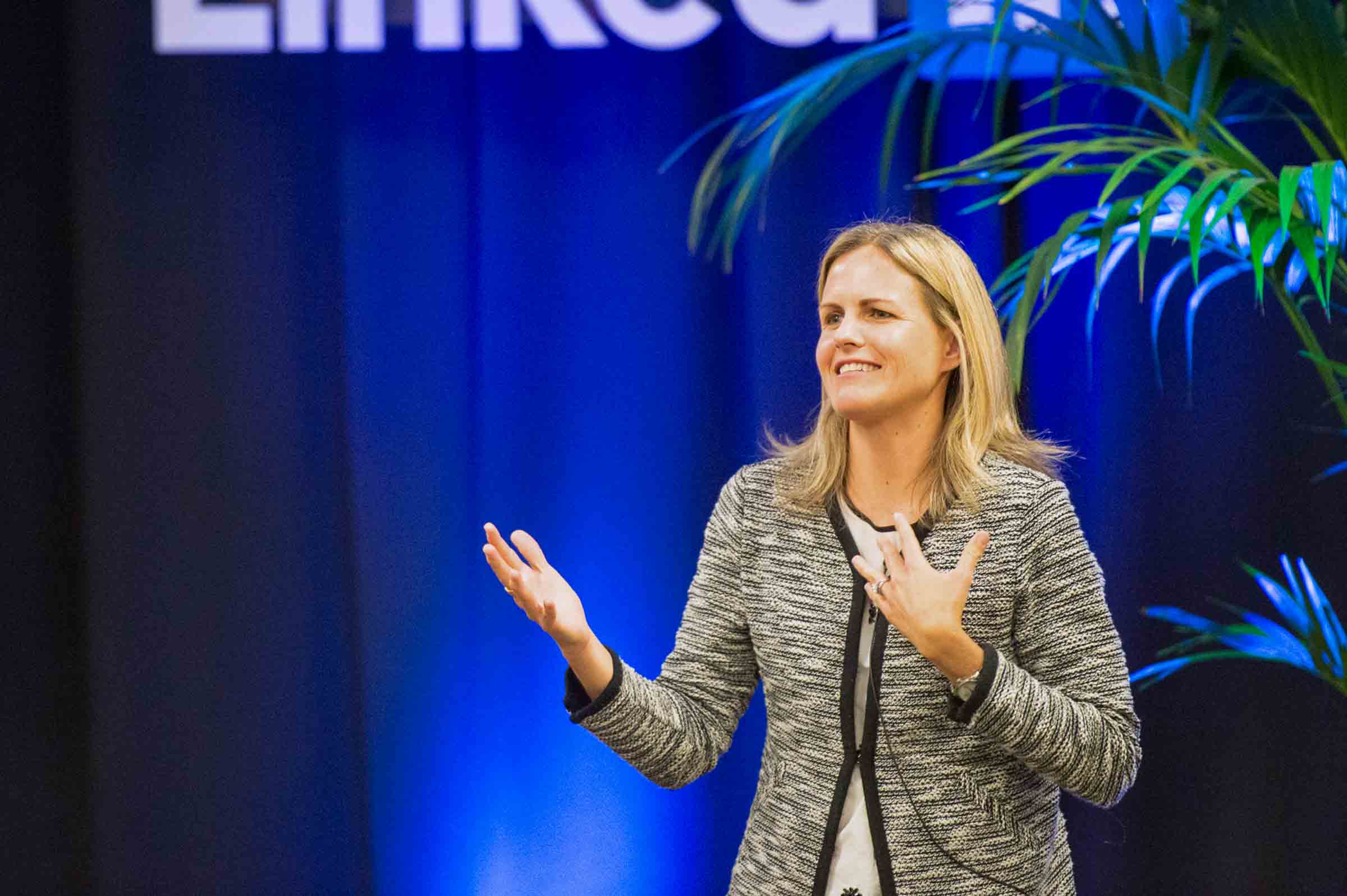 Female executive speaking on stage with animated gestures against blue backdrop with green foliage accents