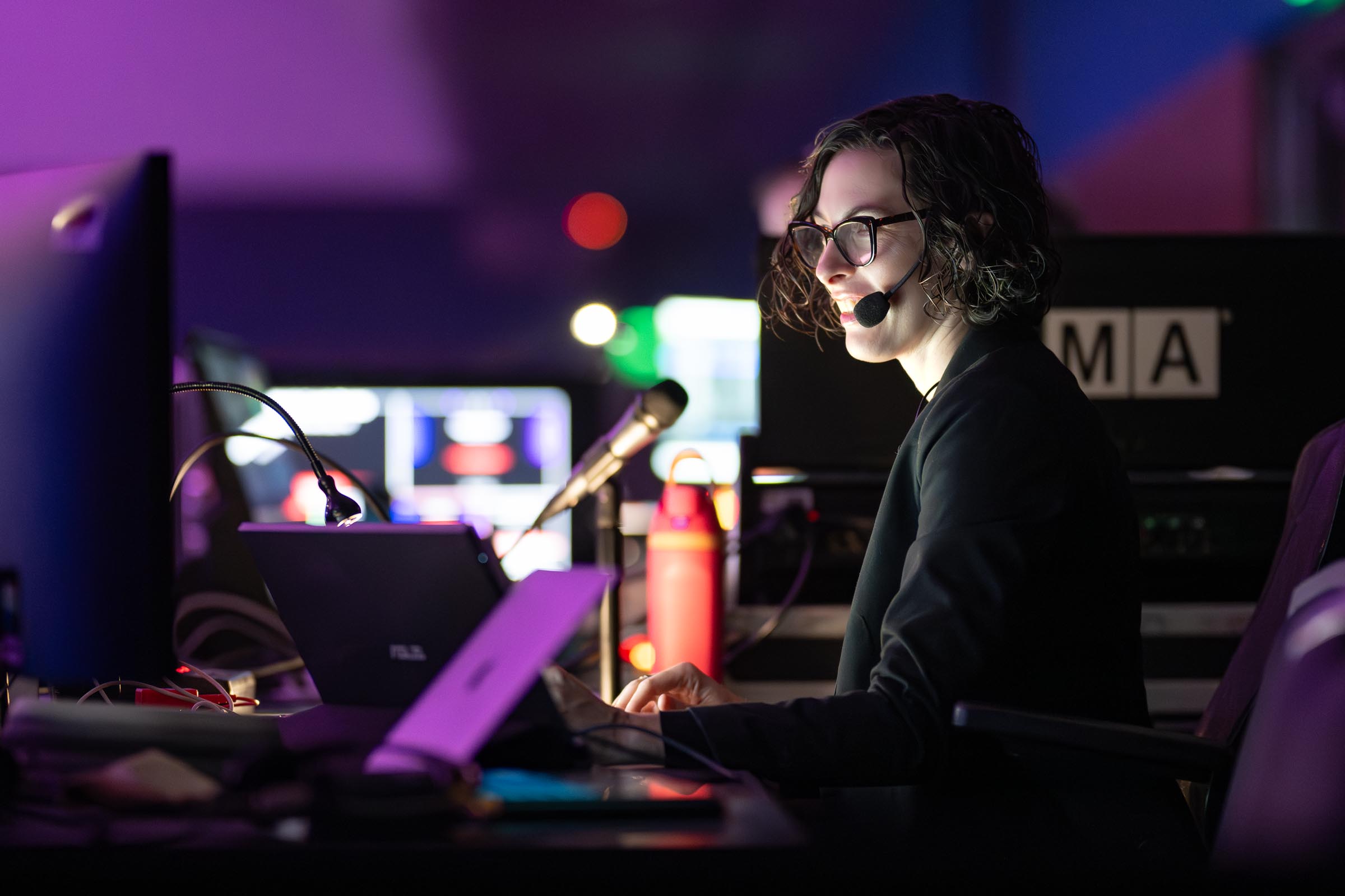 Woman wearing glasses speaking at microphone against vibrant blue and magenta stage lighting