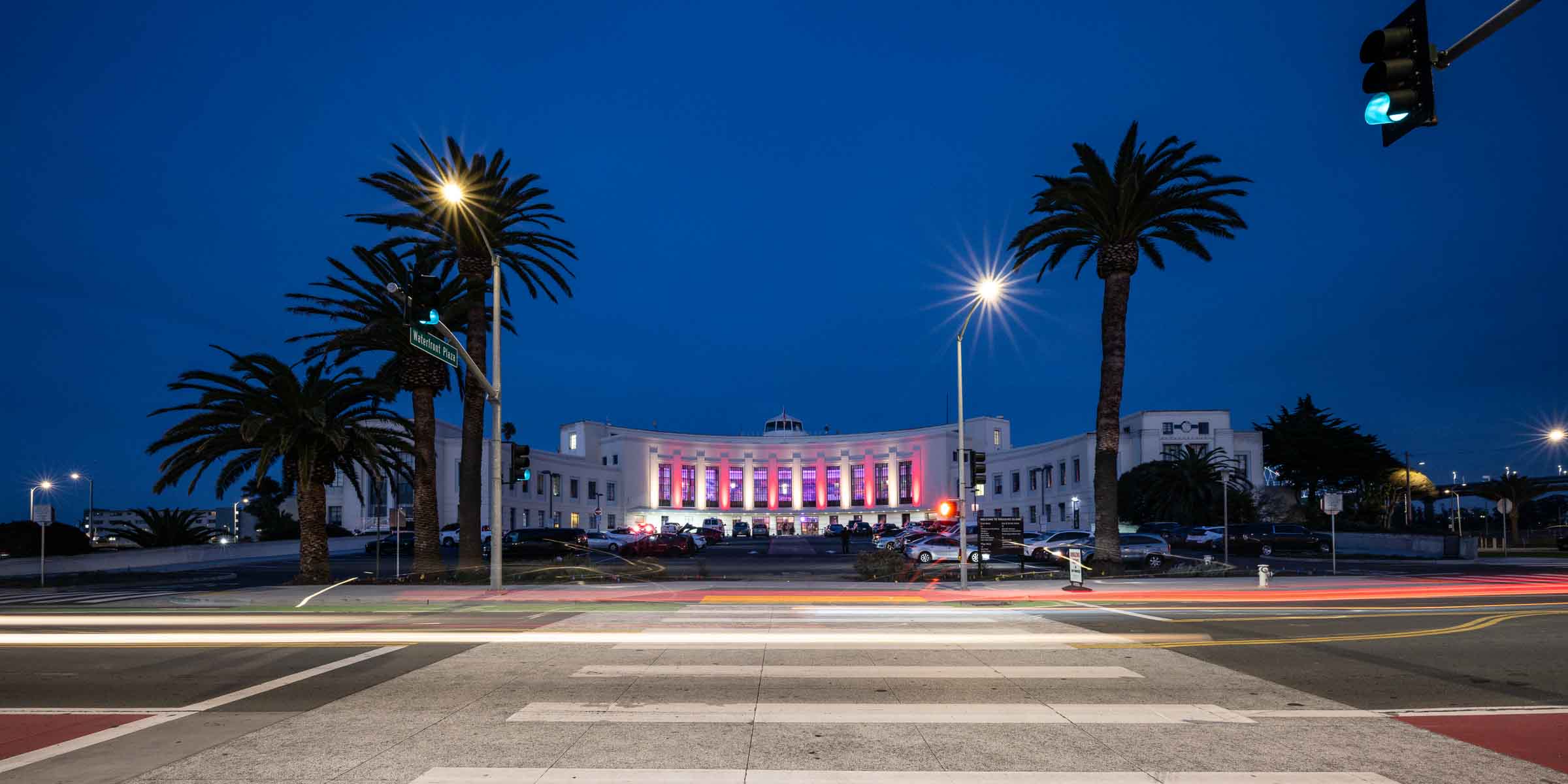 Art Deco event venue with pink-lit windows and palm trees at twilight with long-exposure traffic light trails