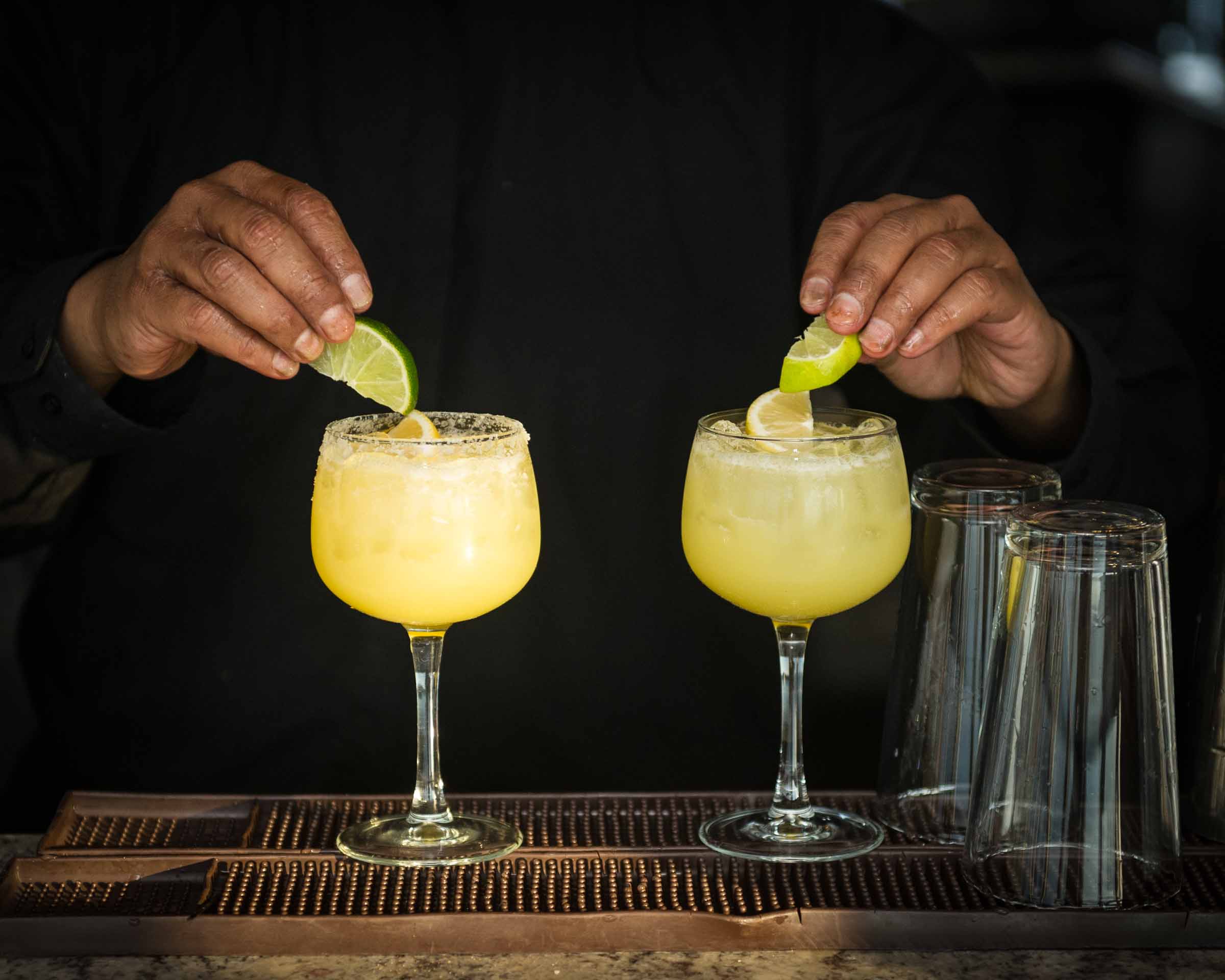 Bartender garnishing golden margarita-style cocktails with lime wedges on copper bar mat